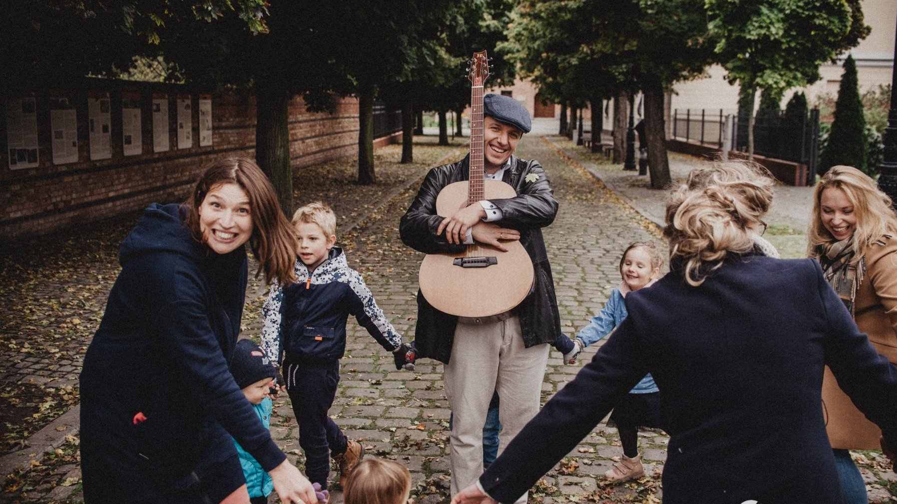 Zdjęcie przedstawia grupę ludzi, którzy tańczą w kółku trzymając się za ręce. W środku znajduje się mężczyzna z gitarą.