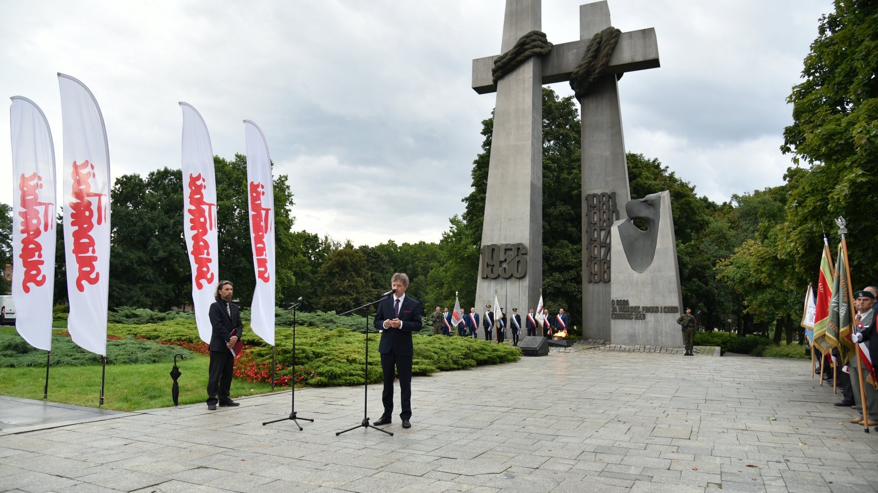 Zdjęcie przedstawia Zdjęcie przedstawia przemawiającego do mikrofonu przewodniczącego wielkopolskiej Solidarności. W tle widać pomnik dwóch krzyży.