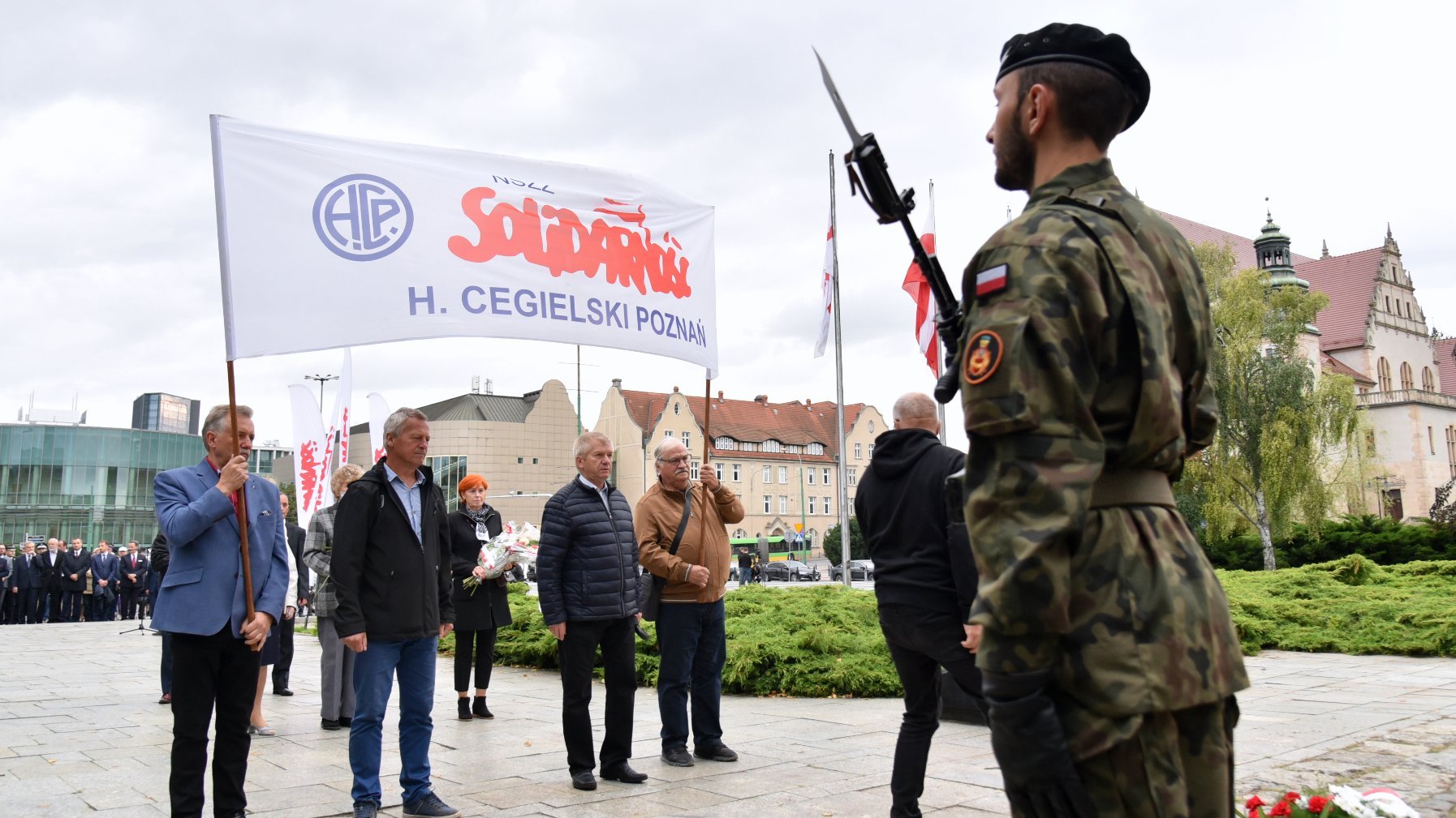 Zdjęcie przedstawia ludzi składających kwiaty, trzymających flagę z napisem "Solidarność".