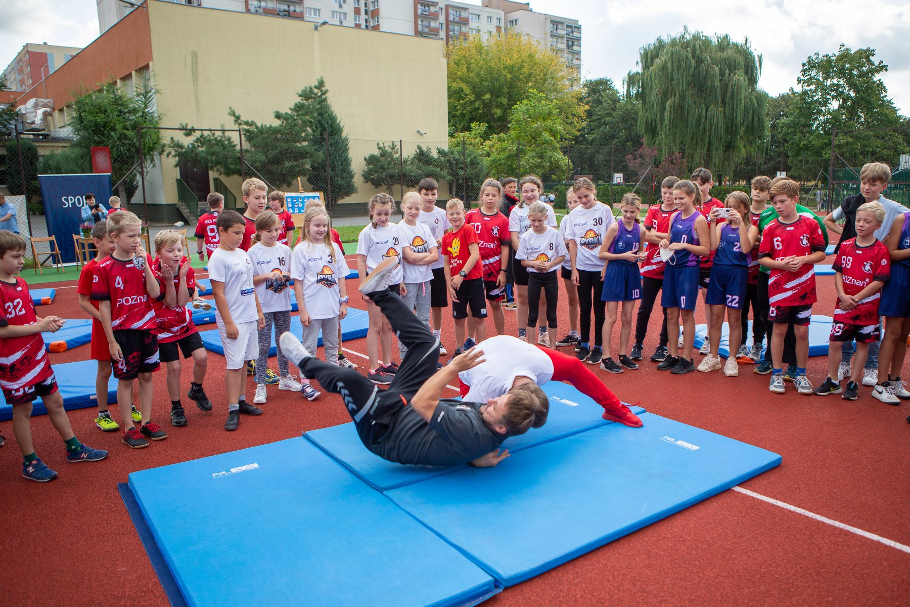 Na zdjęciu boisko szkolne, na macie dwóch sportowców, wokół dzieci - grafika artykułu