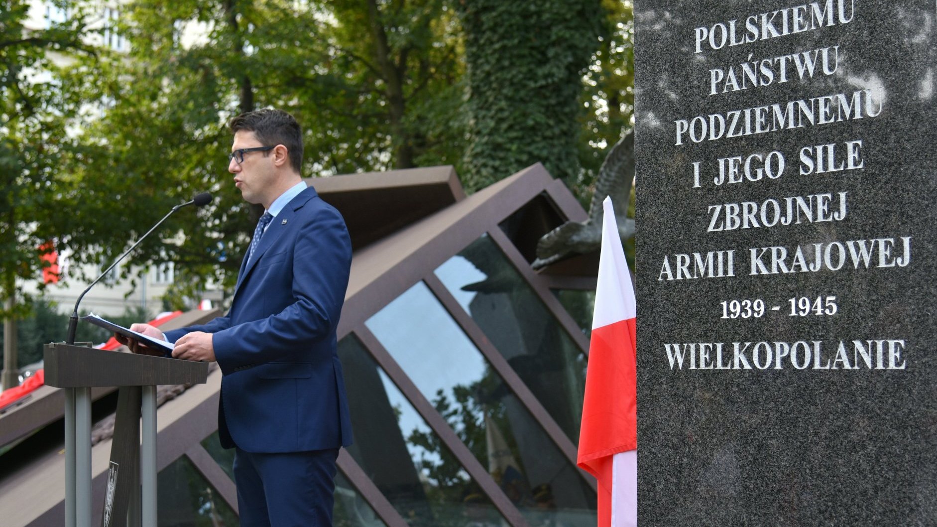 Zdjęcie przedstawia mężczyznę przy mównicy. Na pierwszym planie widać tablicę poświęconą Polskiemu Państwu Podziemnemu.