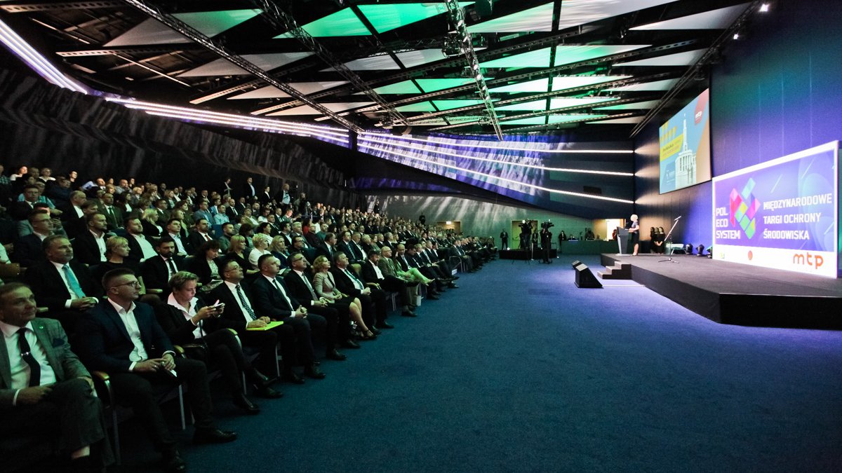 Zdjęcie przedstawia ludzi siedzących na sali konferencyjnych. Na scenie przemawia kobieta.