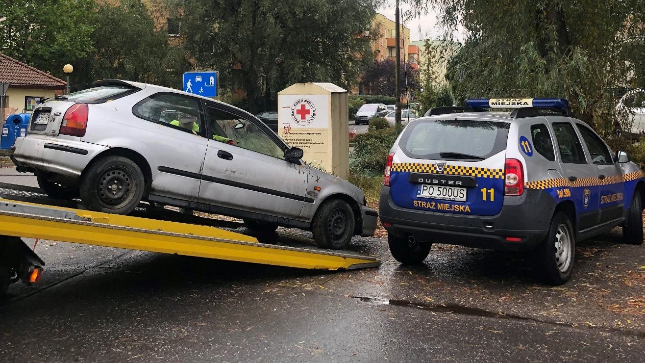 Zdjęcie przedstawia holowany samochód wciągany na lawetę. Obok znajduje się auto straży miejskiej.