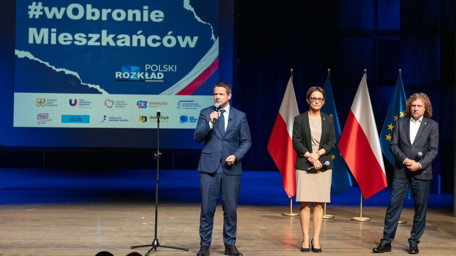 Na zdjęciu trzy osoby na scenie, w tym Rafał Trzaskowski, prezydent Warszawy, mówi do mikrofonu. W tle plansza z napisem: #wObronie Mieszkańców/ Polski Rozkład