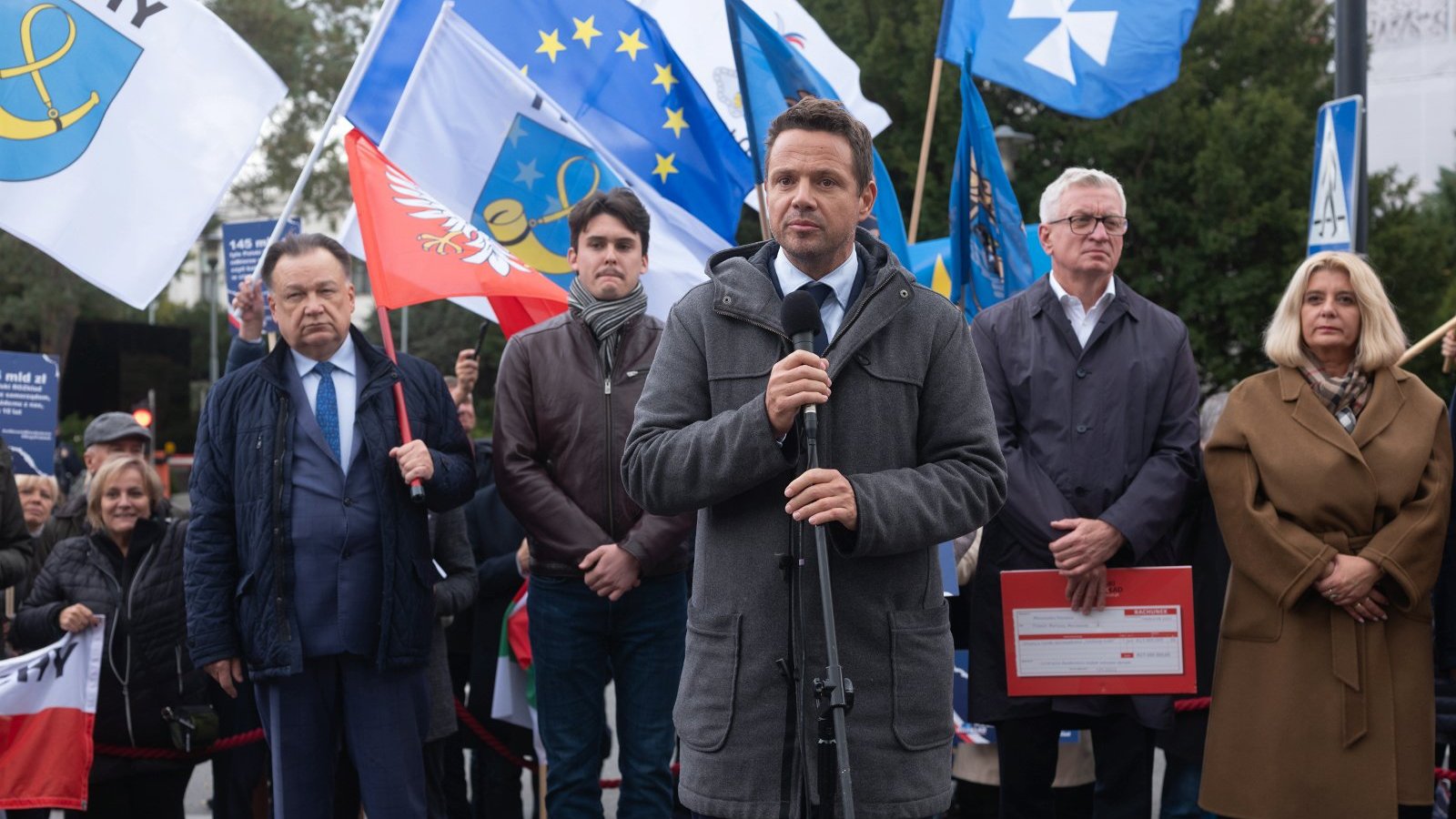 Na zdjęciu samorządowcy z flagami swoich miejscowości, na pierwszym planie, przy mikrofonie, Rafał Trzaskowski, za nim Jacek Jaśkowiak