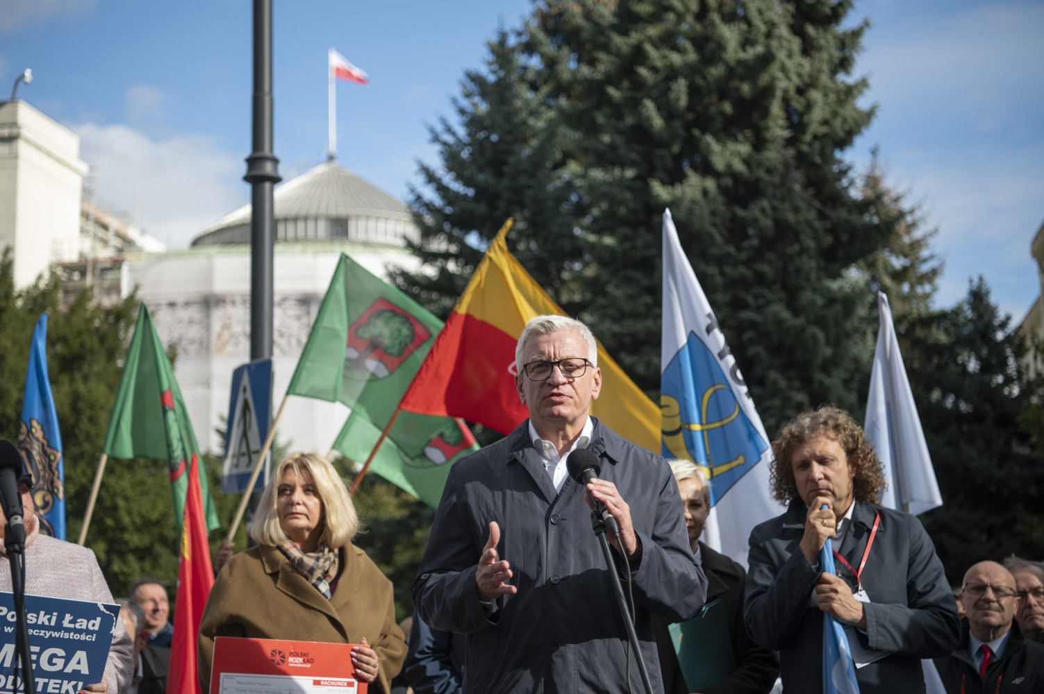 Na zdjęciu Jacek Jaskowiak, prezydent Poznania, przy mikrofonie, za nim inni samorządowcy z flagami, w tle budynek Sejmu - grafika artykułu