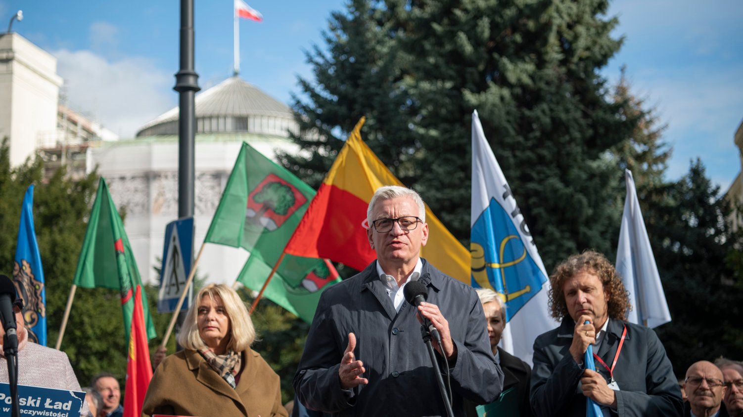 Na zdjęciu Jacek Jaskowiak, prezydent Poznania, przy mikrofonie, za nim inni samorządowcy z flagami, w tle budynek Sejmu