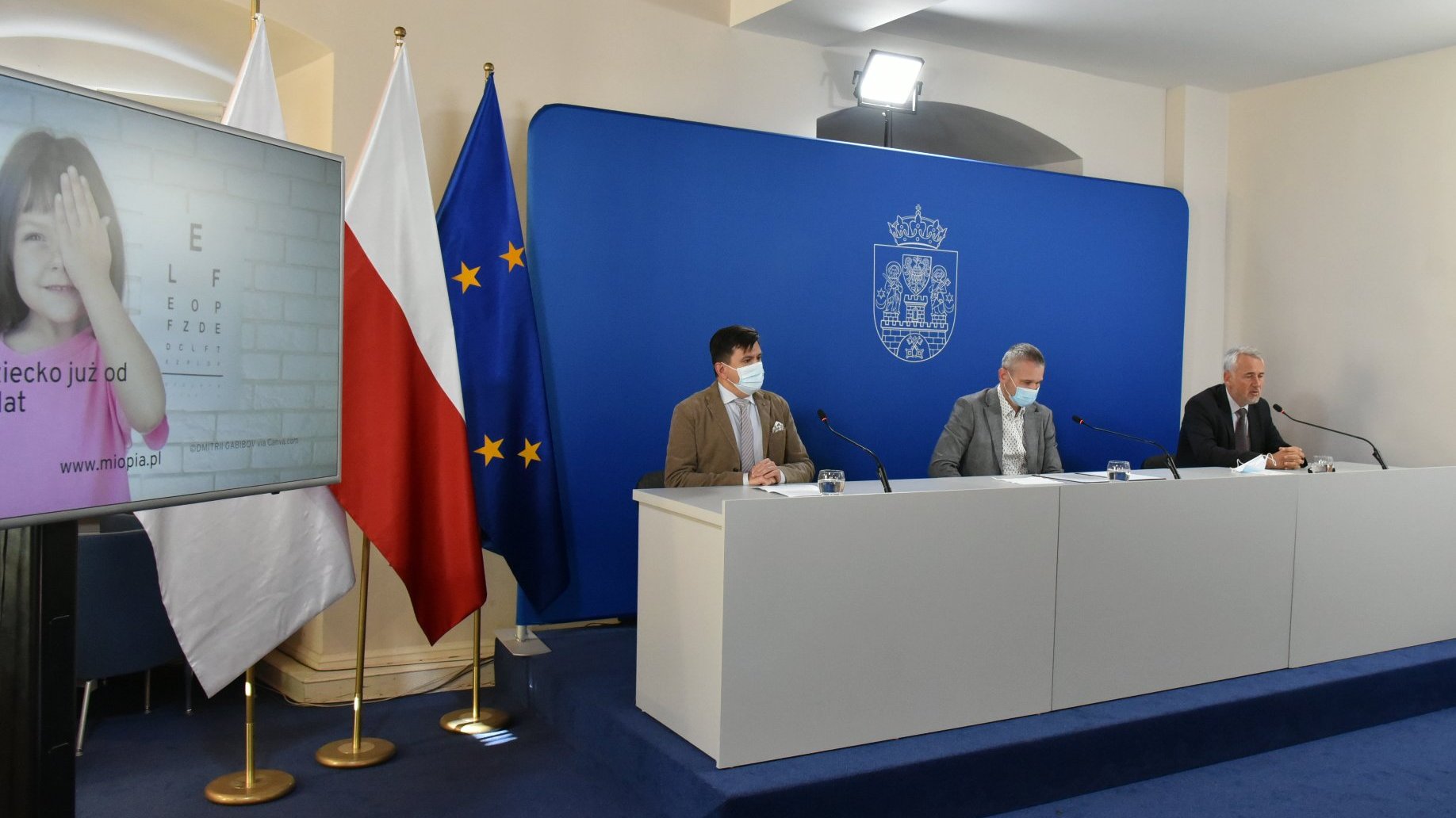 Na zdjęciu sala konferencyjna, za stołem siedzi 3 mężczyzn, na pierwszym planie telewizor, w kadrze dziewczynka zasłaniająca sobie jedno oko