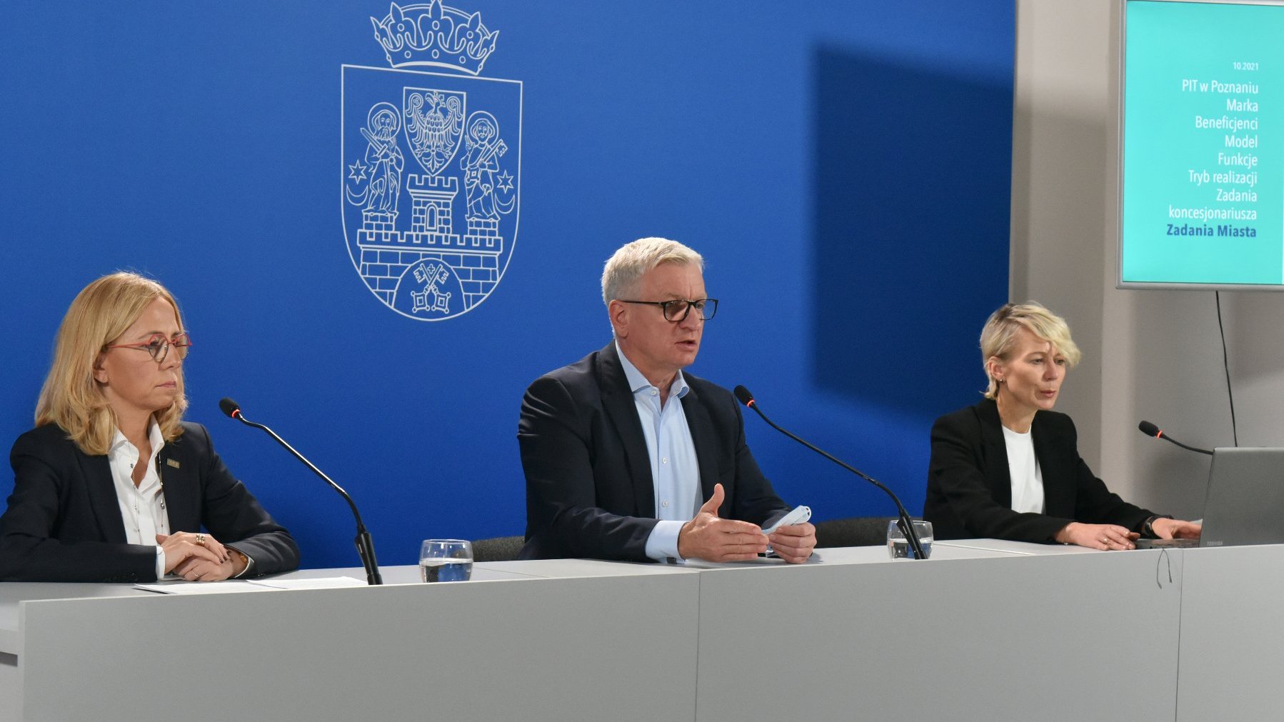 Na zdjęciu trzy osoby za stołem konferencyjnym, w środku Jacek Jaśkowiak, prezydent Poznania, za nimi granatowa ścianka z herbem miasta