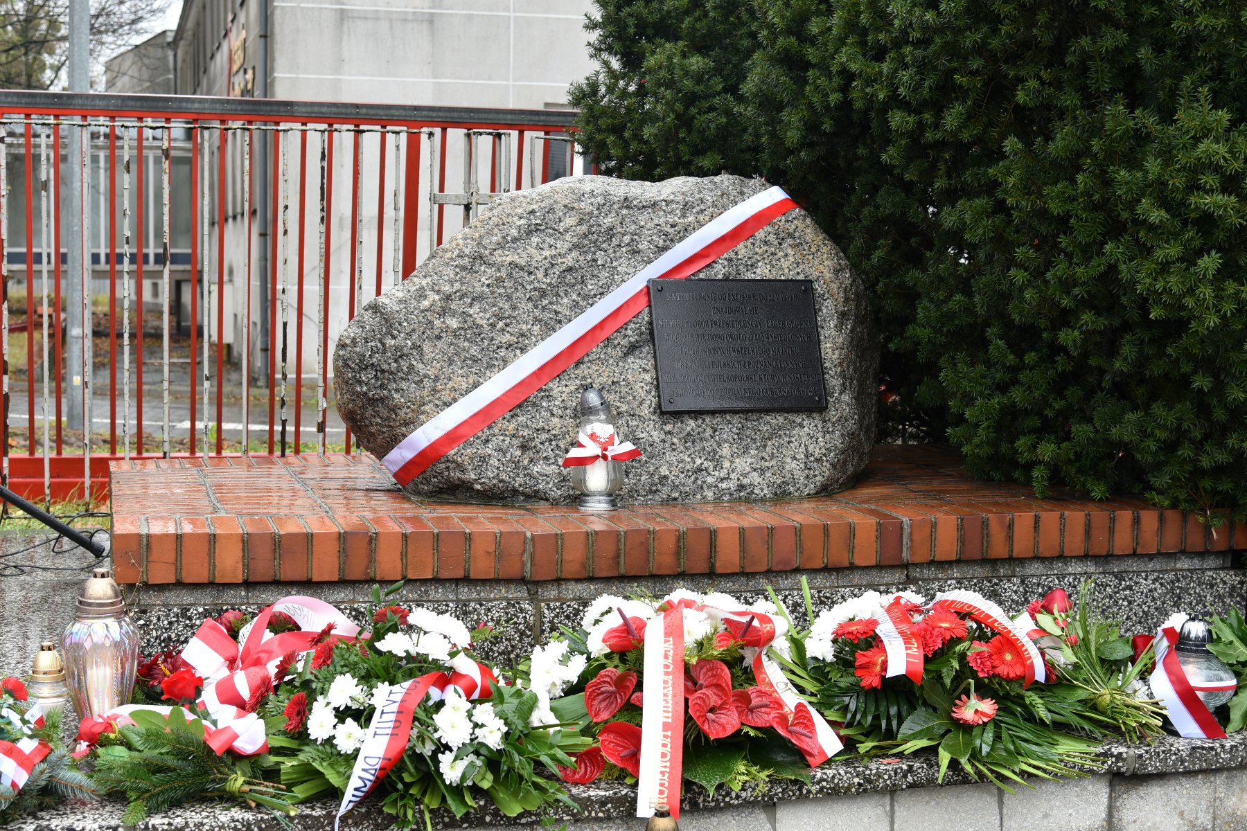 Zdjęcie przedstawia głaz upamiętniający miejsce, gdzie od końca października 1939 r. do maja 1940 r. działał niemiecki obóz przesiedleńczy. Jest on przepasany biało czerwoną wstęgą, pod nim znajdują się kwiaty. - grafika artykułu