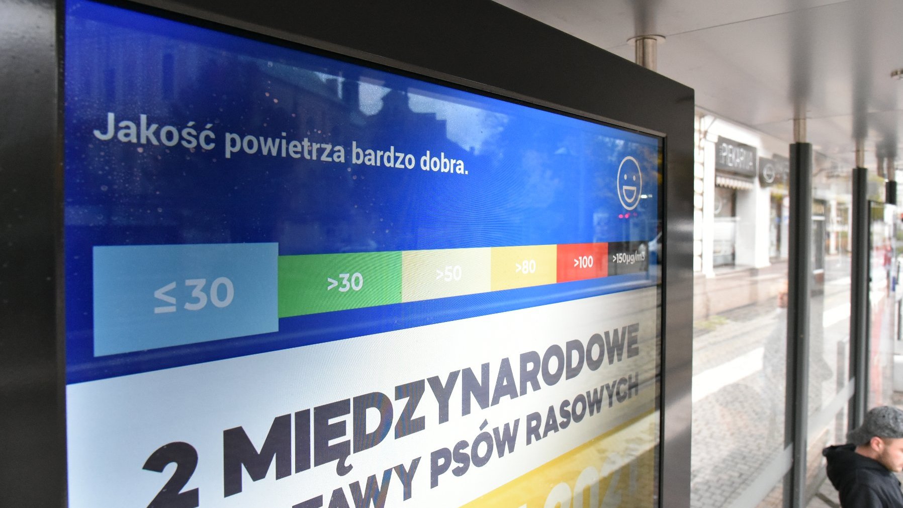 Galeria zdjęć przedstawia ekran z reklamą i informacją o jakości powietrza, znajdujący się na przystanku komunikacji miejskiej.