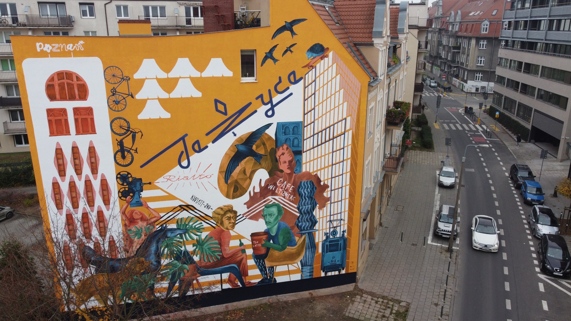 Galeria zdjęć przedstawia nowy mural na Jeżycach. Widać na nim m.in. dwóch poetów Słowackiego i Mickiewicza. Obok znajduje się ulica z samochodami.