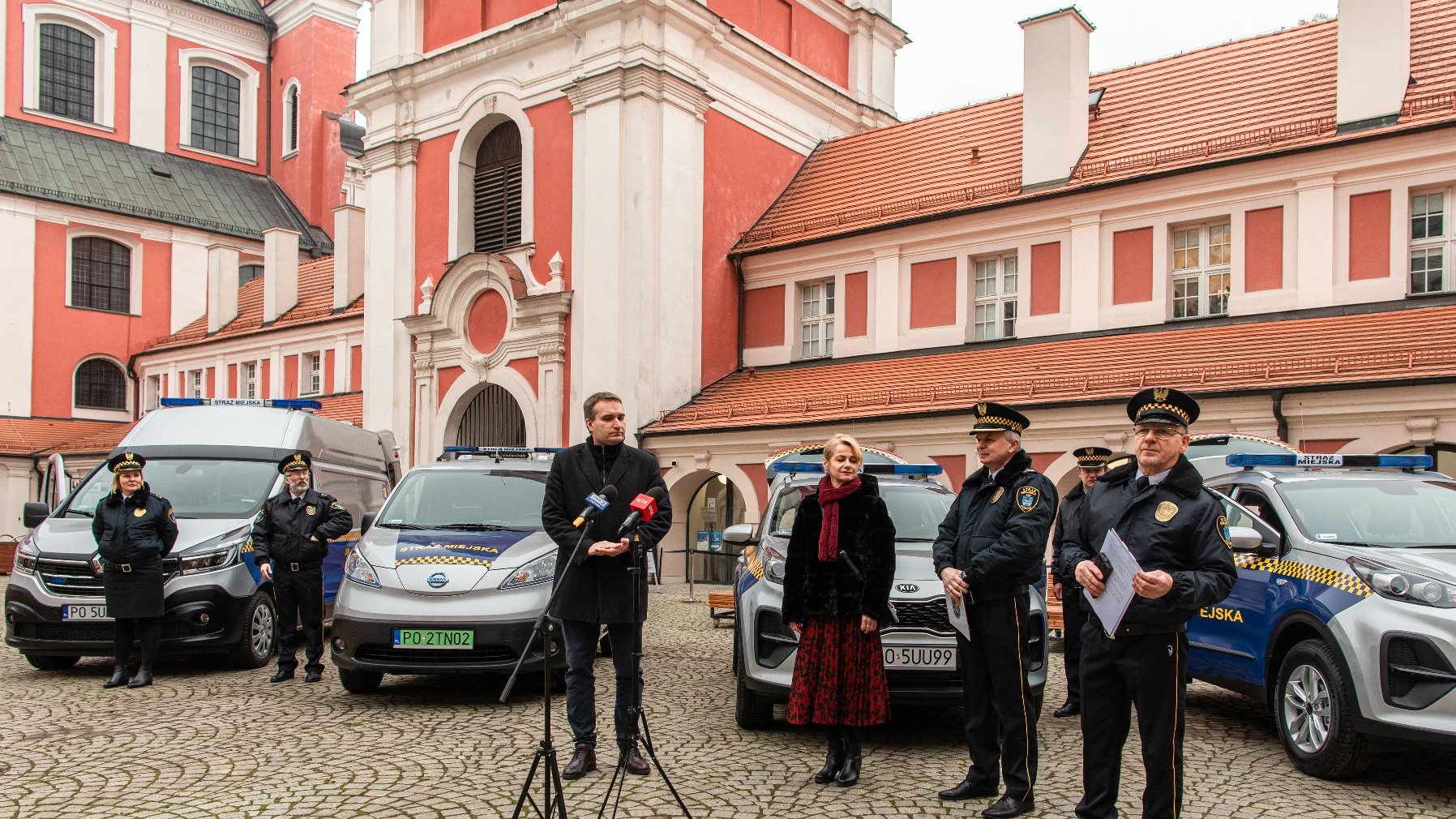 Galeria zdjęć przedstawia uczestników konferencji prasowej na dziedzińcu urzędu miasta. W tle widać nowe samochody.