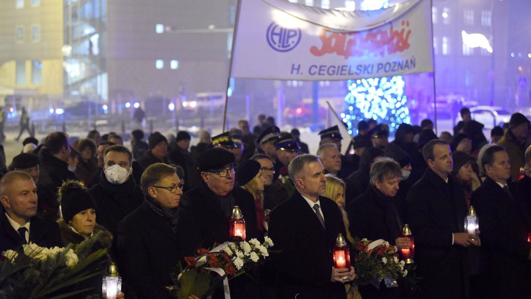 Grupa osób na pl. Mickiewicza, w dłoniach trzymają znicze. Na drugim planie flaga ZSZZ "Solidarność" H. Cegielski.