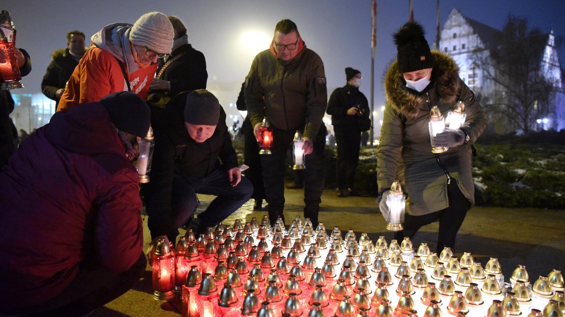 Grupa osób układa we flagę Polski białe i czerwone znicze.