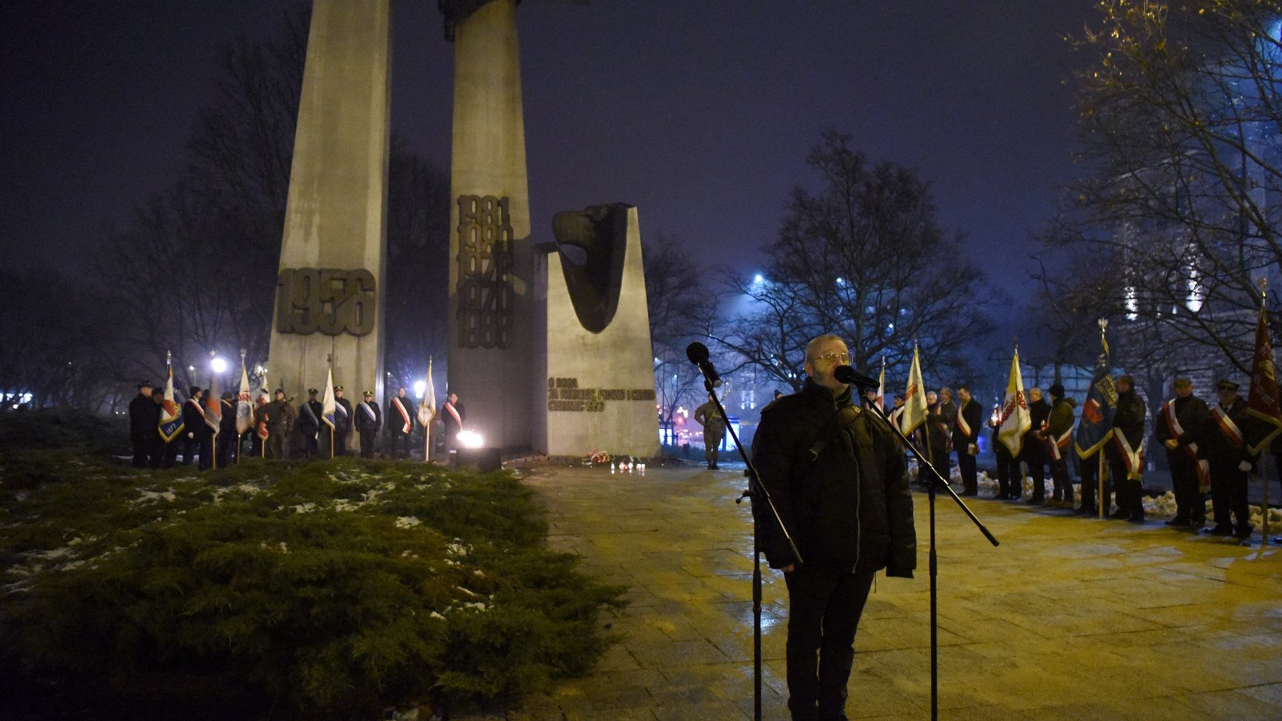 Osoba przemawiająca do mikrofonu. W tle pomnik Poznańskiego Czerwca w kształcie dwóch wielkich krzyży.