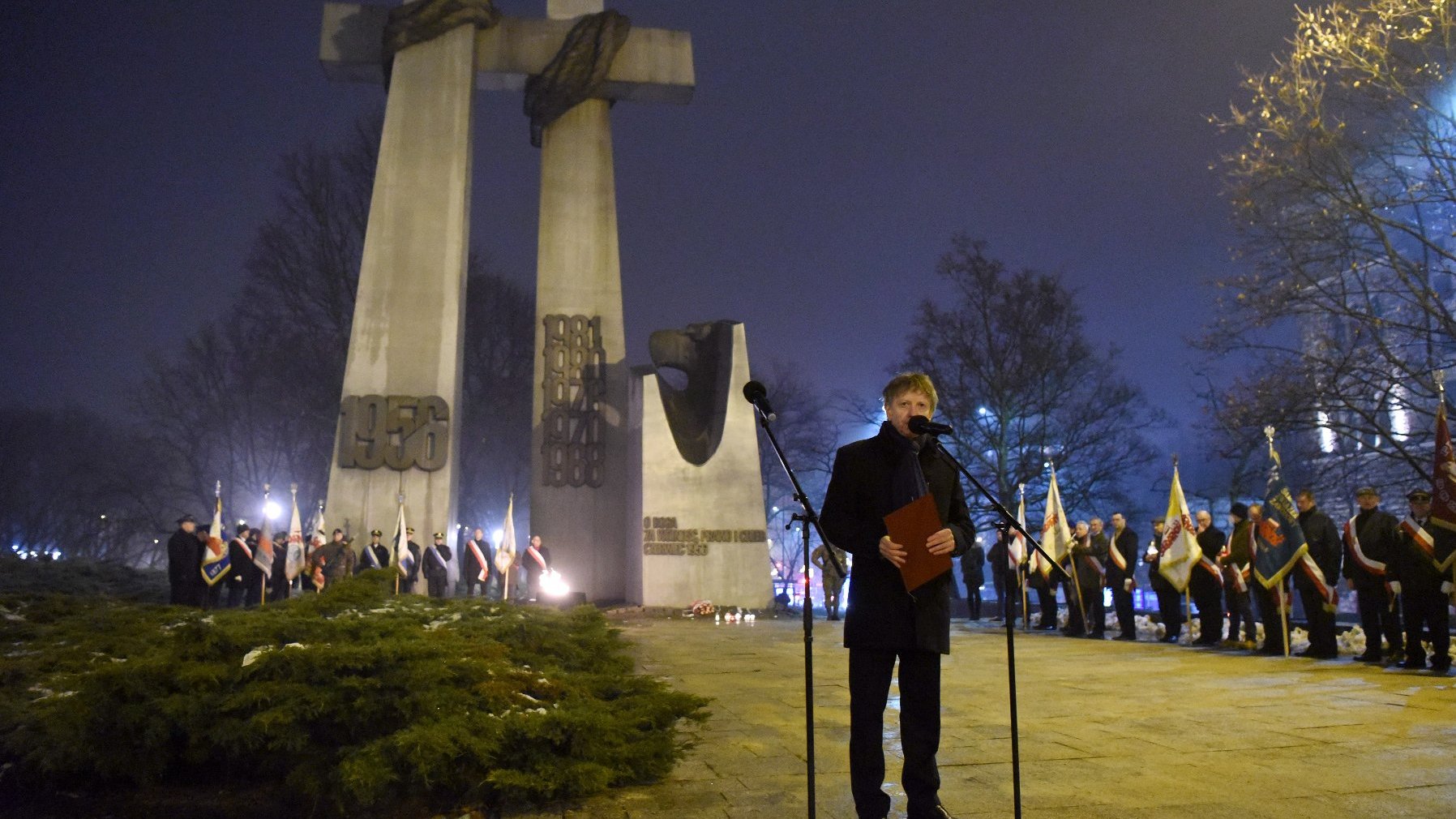 Osoba przemawiająca do mikrofonu. W tle pomnik Poznańskiego Czerwca w kształcie dwóch wielkich krzyży.