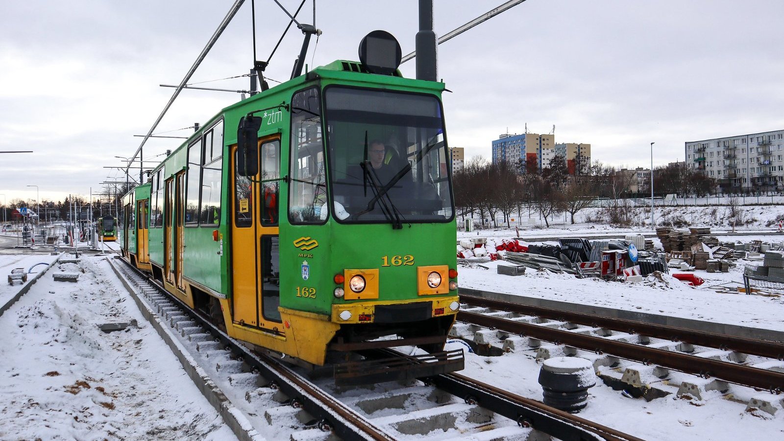 Tramwaj stojący na torowisku, w tle bloki, na trawie śnieg