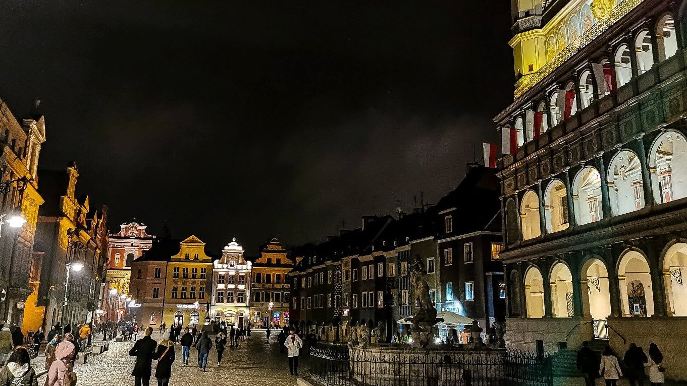 Zdjęcie przedstawia Stary Rynek po zmroku. Widać na nim budynki i spacerujących ludzi.