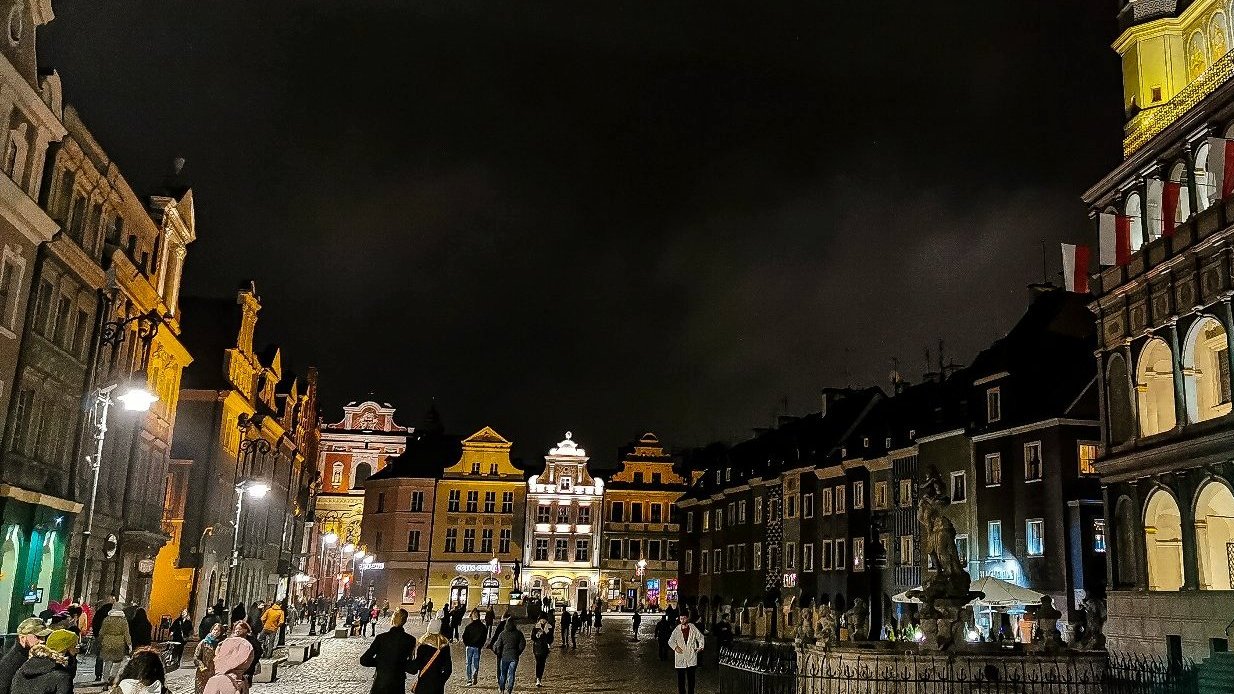 Zdjęcie przedstawia Stary Rynek po zmroku. Widać na nim budynki i spacerujących ludzi.