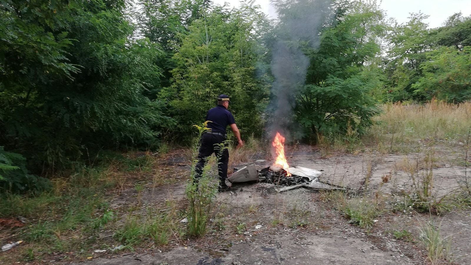 Zdjęcie przedstawia strażnika miejskiego gaszącego palące się odpady. Obok znajdują się drzewa.