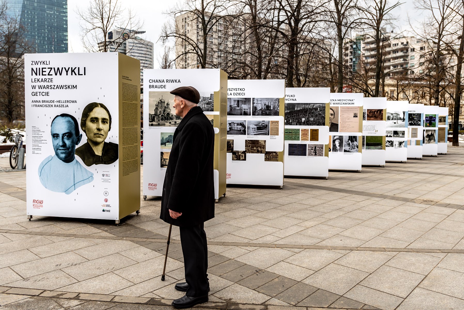 Na zdjęciu wystawa na świeżym powietrzu, kilkanaście stojaków, przygląda im się starszy mężczyzna w ciemnym płaszczu - grafika artykułu