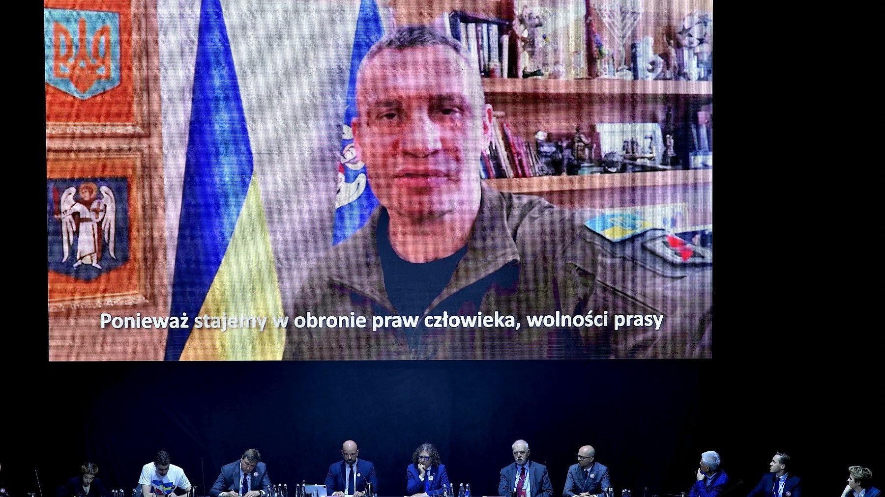 Zdjęcie przedstawia uczestników obrad siedzących przy stole oraz telebim, na którym widać Witalija Kliczko, mera Kijowa.