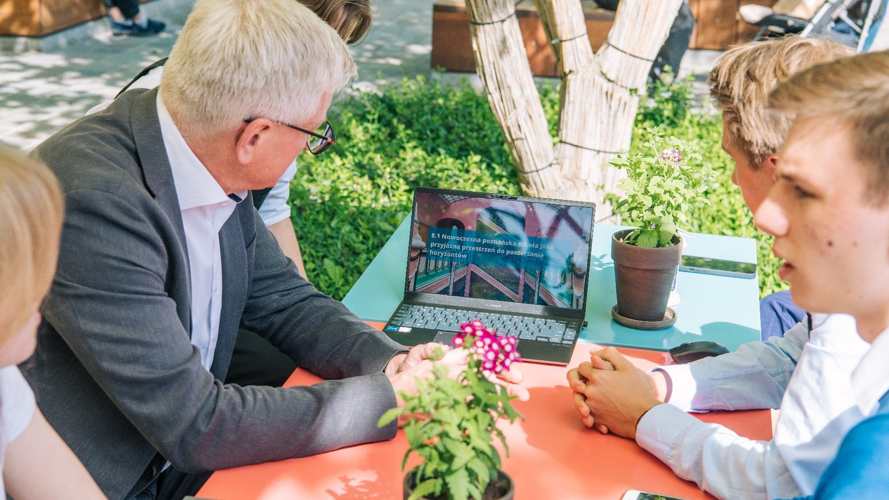 Jacek Jaśkowiak i młodzi ludzie siedzą w ogródku kawiarnianym, na stoliku leży laptop z prezentacją, wszyscy patrzą na niego