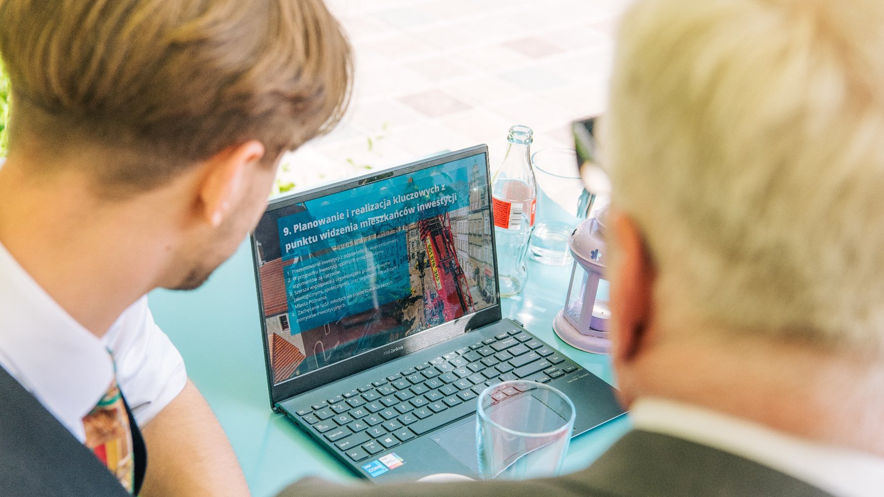 Na zdjęciu widać głowy dwóch mężczyzn, patrzących na laptop, którzy leży przed nimi na kawiarnianym stoliku, na ekranie prezentacja