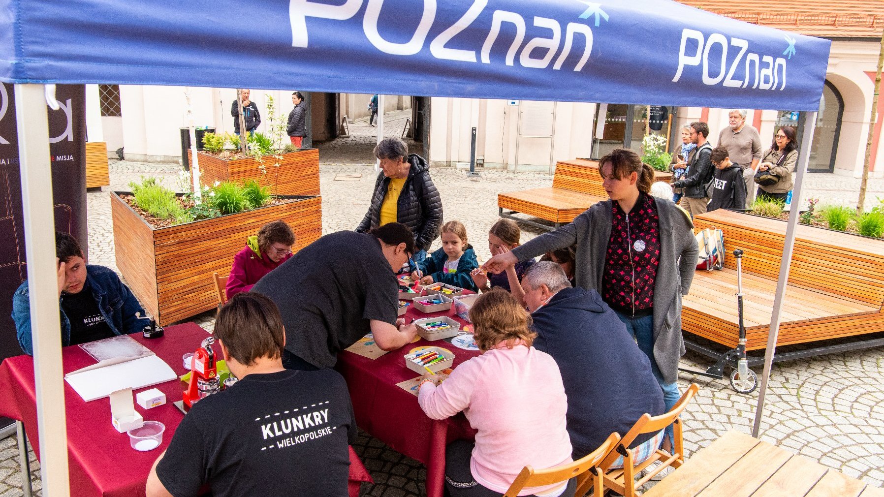 Zdjęcie przedstawia stoły, na których leżą przybory plastyczne oraz siedzących przy nich ludzi, głównie dzieci.