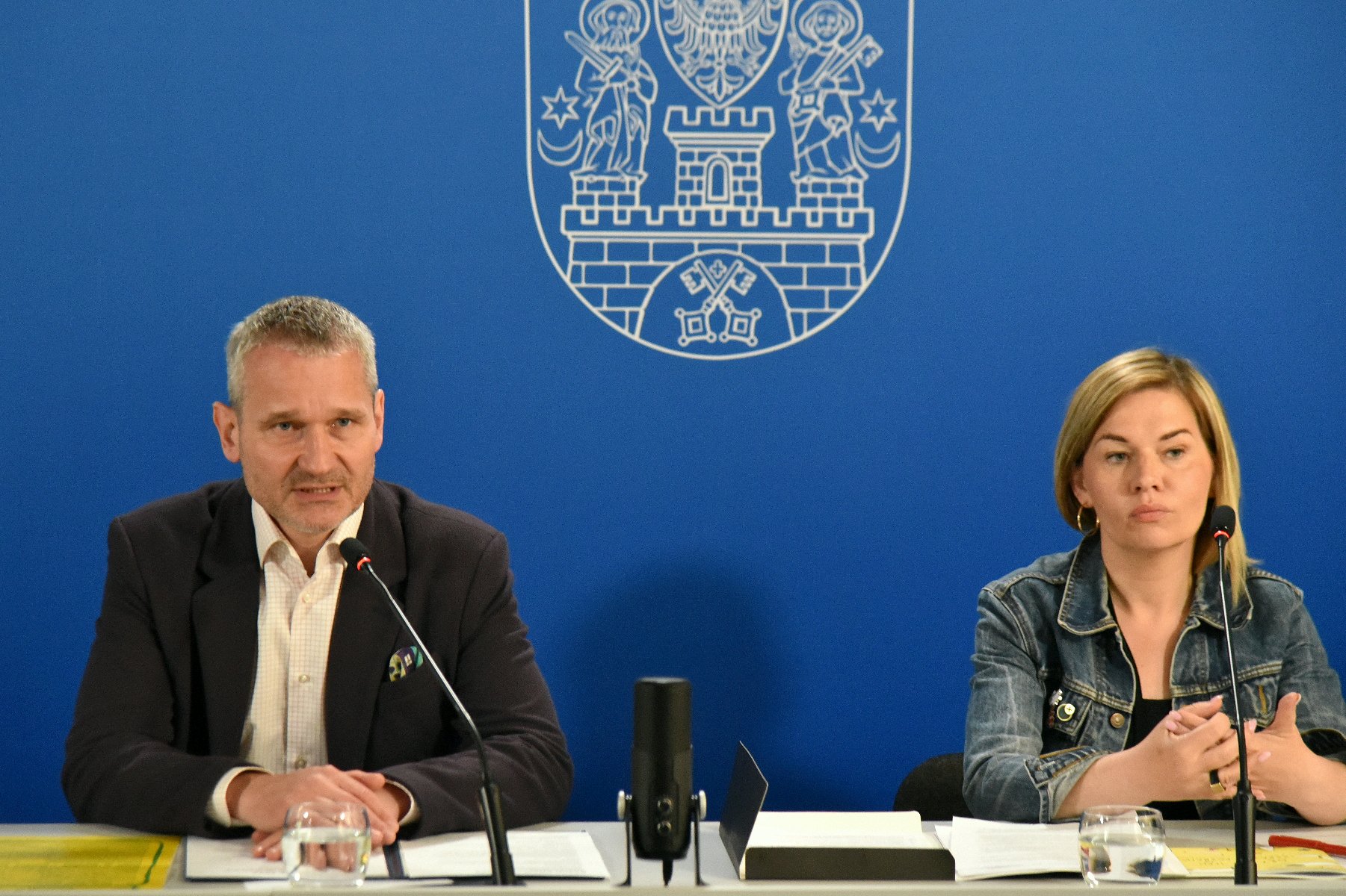 Na zdjęciu mężczyzna i kobieta za stołem konferencyjnym, za nimi gratatowa ścianka z herbem miasta - grafika artykułu