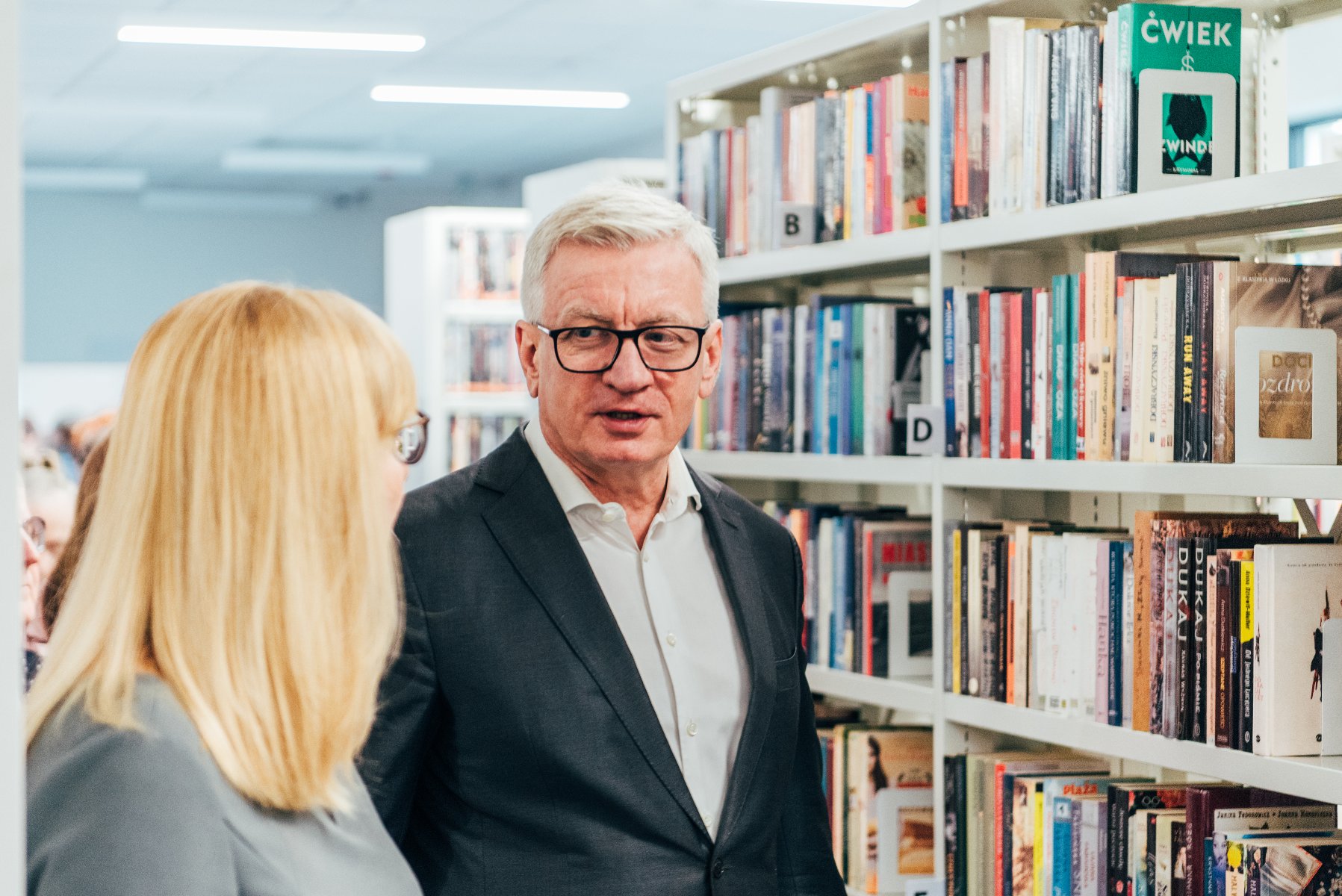 Na zdjęciu prezydent Poznania rozmawia z dyrektorką biblioteki (tyłem), w tle regał z książkami - grafika artykułu