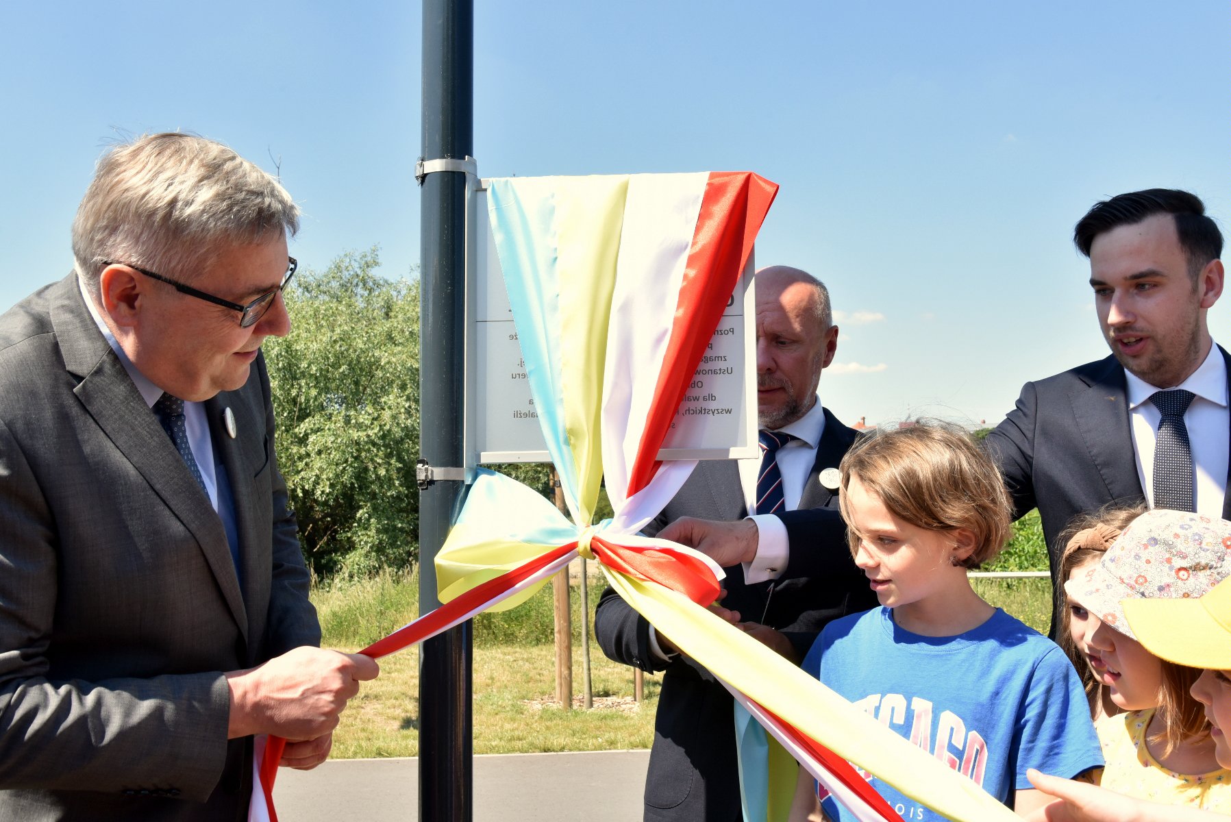 Na zdjęciu kilka osób - trzej dorośli mężczyźni oraz kilkoro dzieci - odsłaniają tabliczkę z nazwą skweru, ciągnąc za wstęgi w kolorach flag - grafika artykułu