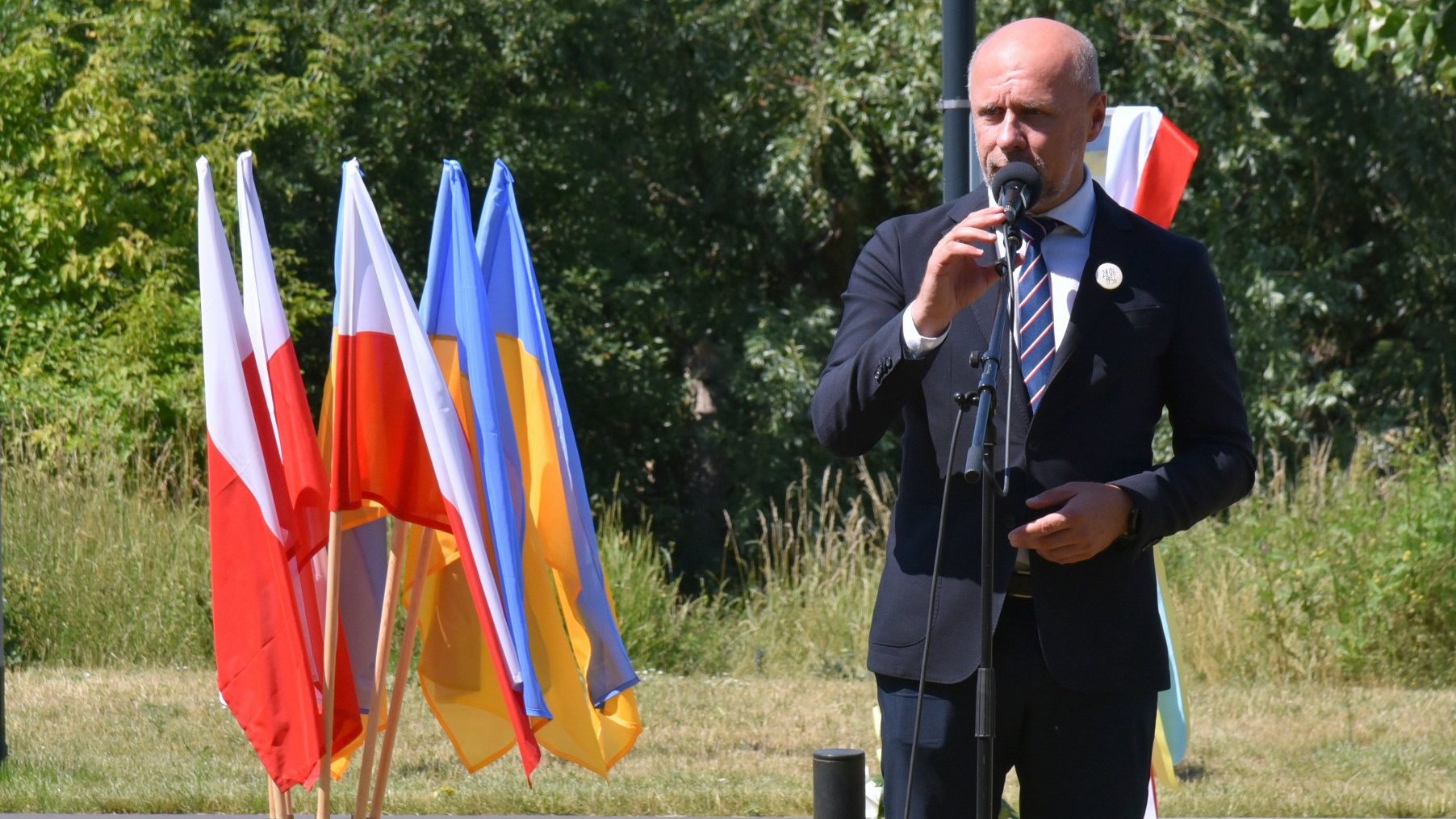 Zastępca prezydenta Poznania przy mikrofonie, w tle drzewa, obok flagi polskie i ukraińskie