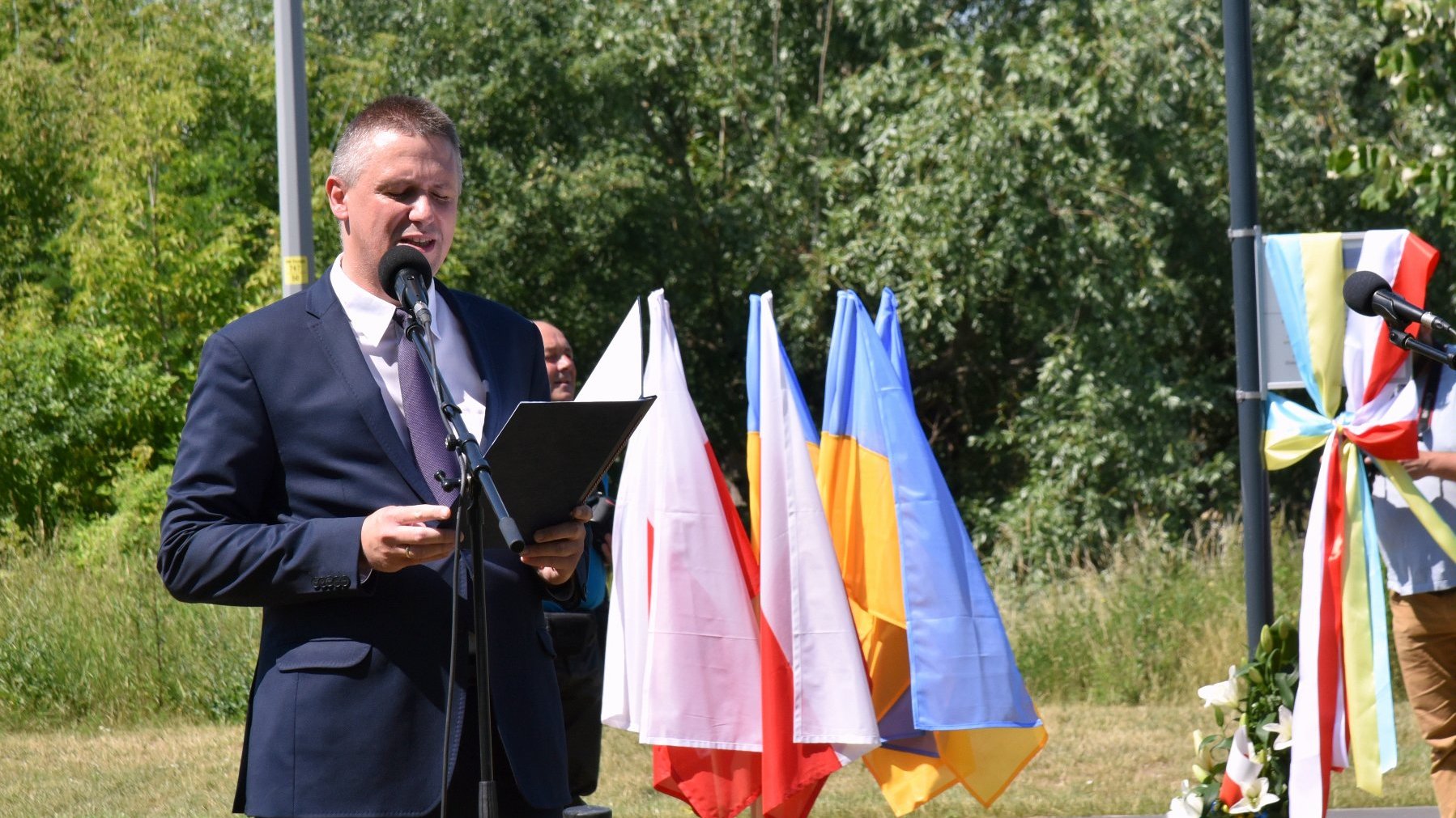 Na zdjęciu dyrektor Gabinetu Prezydenta przy mikrofonie, w tle drzewa i flagi polskie i ukraińskie