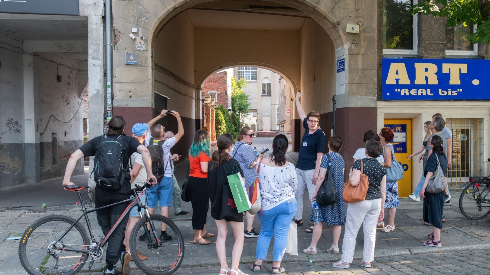 Grupa osób stoi przed budynkiem. Jedna z nich wskazuje ręką w stronę bramy.