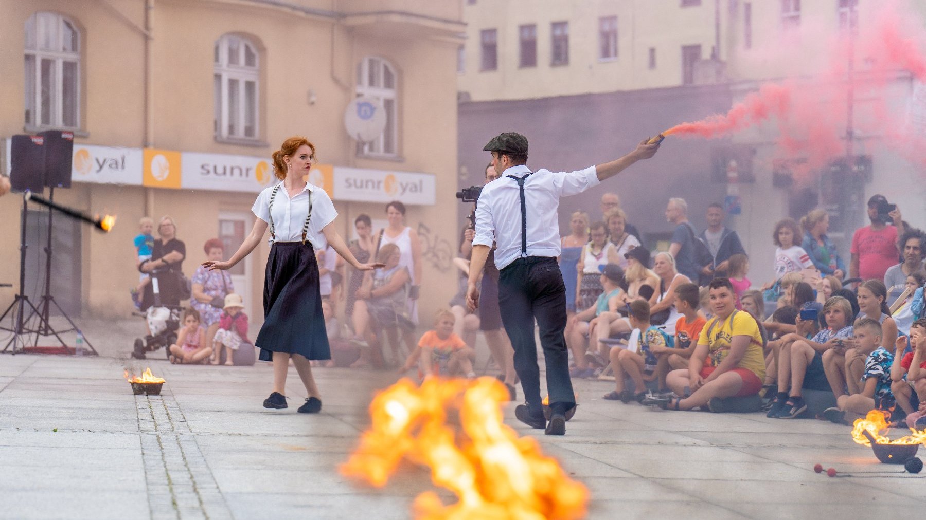 Na zdjęciu dwoje artystów, mężczyzna trzyma kolorową racę, kobieta odrzuca pochodnie, przypatruje im się publiczność