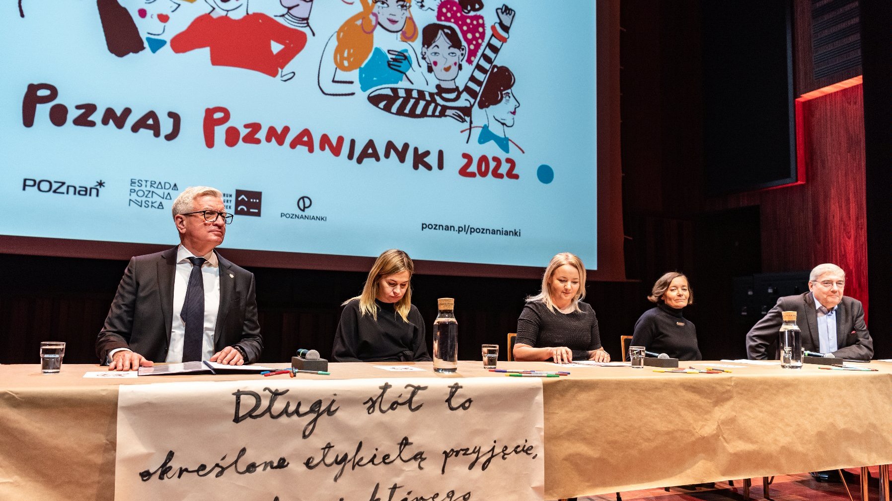 Na zdjęciu pięć osób za stołem, wśród nich prezydent Poznania, w tle ekran z rysunkiem kobiet i napisem: poznaj poznanianki 2022