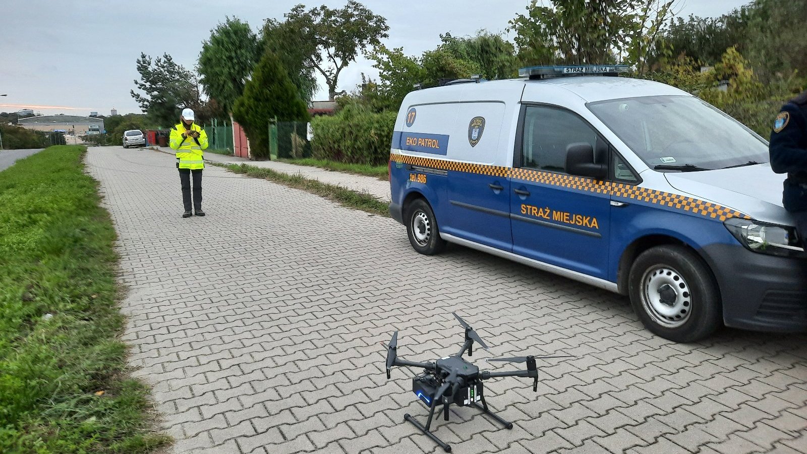 Na zdjęciu dron stojący na ziemi obok samochodu straży miejskiej