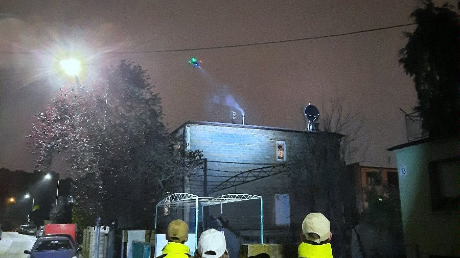 Na zdjęciu dron latający po zmroku nad domami, przed posesjami znajdują się obserwujący ludzie oraz operator drona