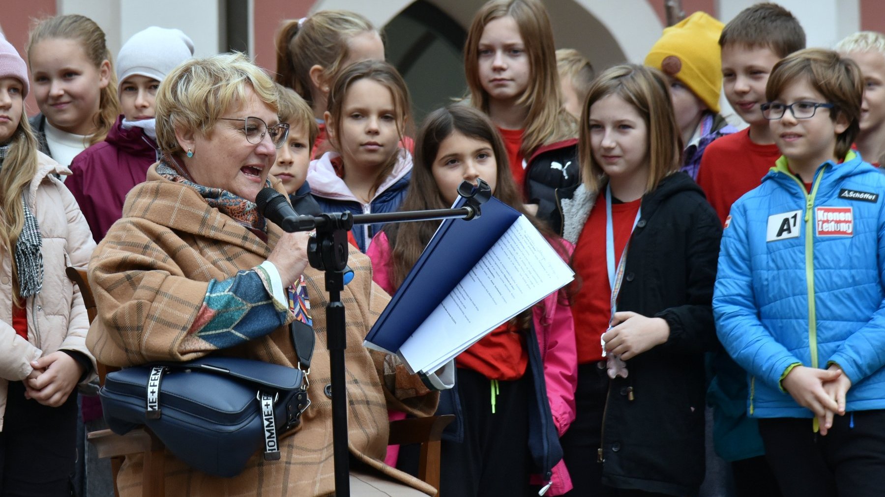 Na zdjęciu kobieta w okularach, czytająca dzieciom, za nią uczniowie