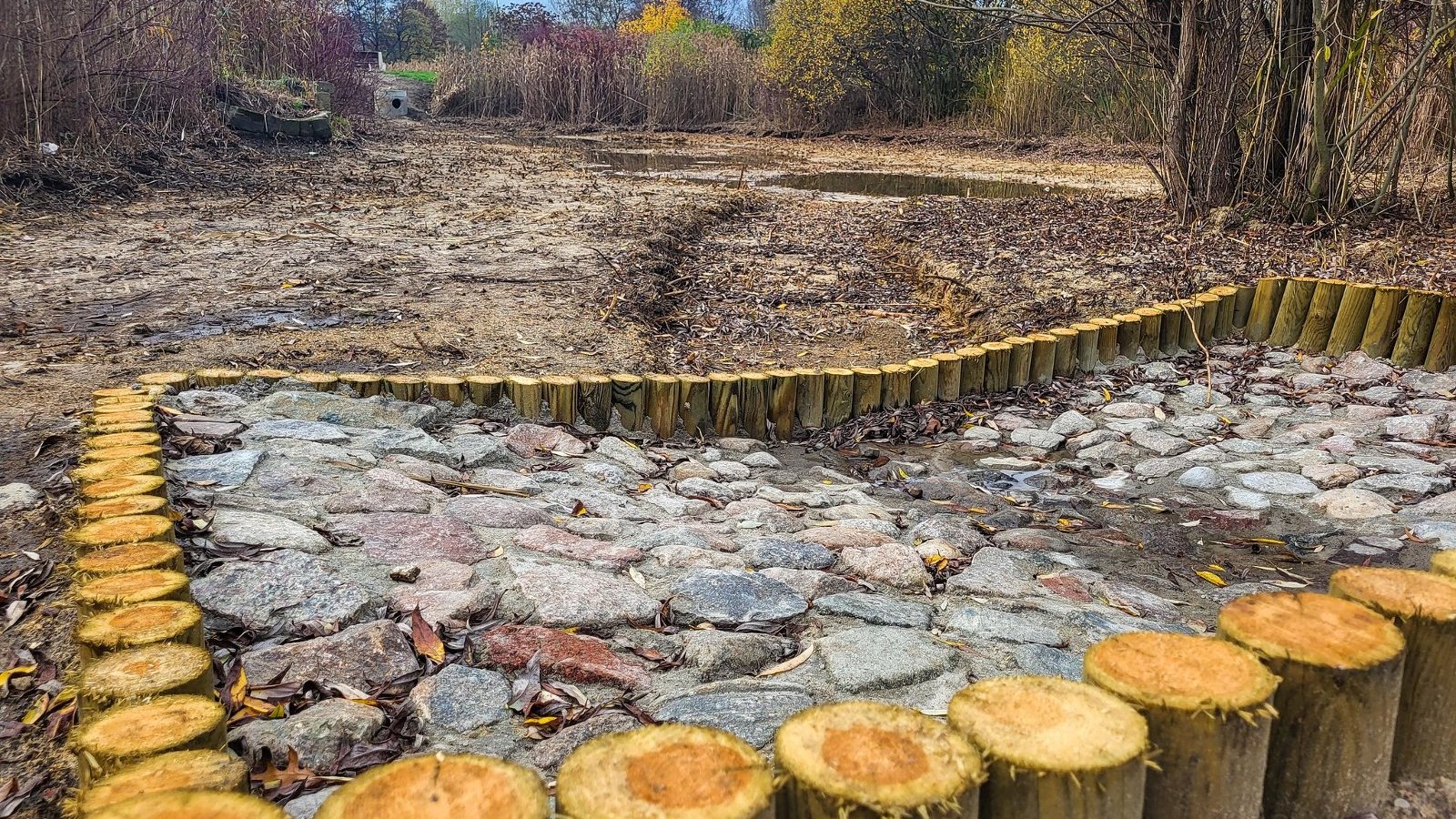 Na zdjęciu konstrukcja z pni i kamieni, w tle staw oraz rośliny