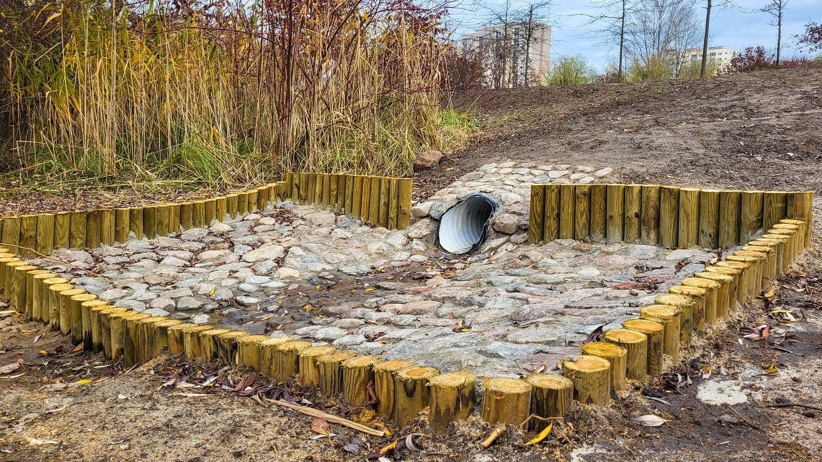 Na zdjęciu konstrukcja z pni i kamieni, w tle wylot rury, na dalszym planie bloki