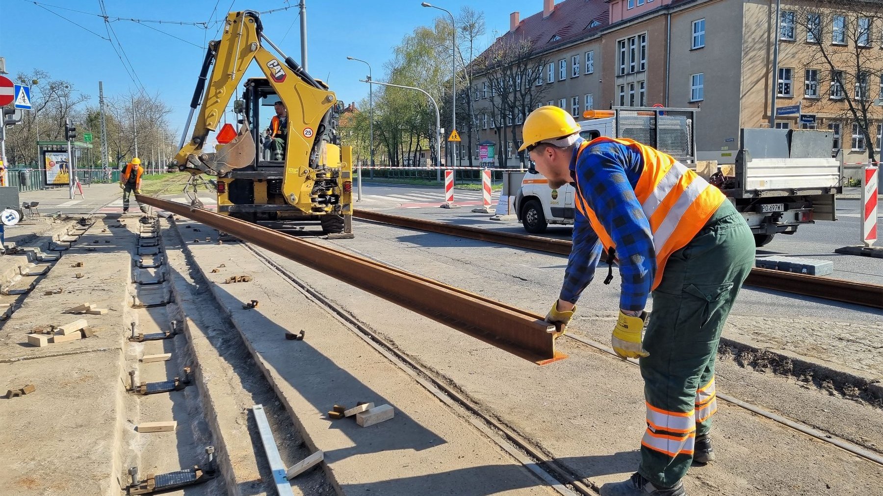 Zdjęcie przedstawia pracowników służb torowych układającyh torowisko. W tle koparka.