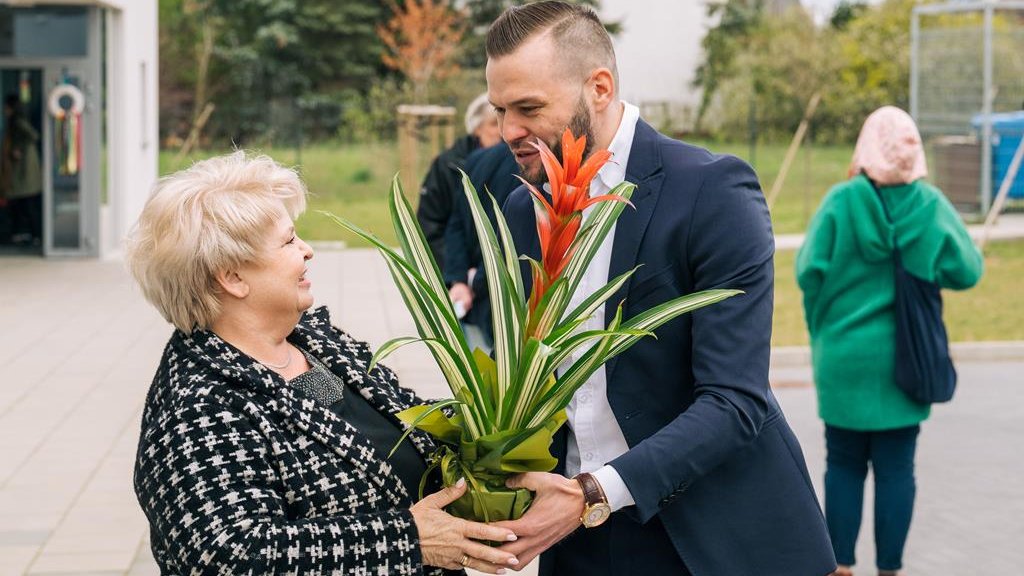 Na zdjęciu mężczyzna w garniturze wręczający kwiat doniczkowy kobiecie