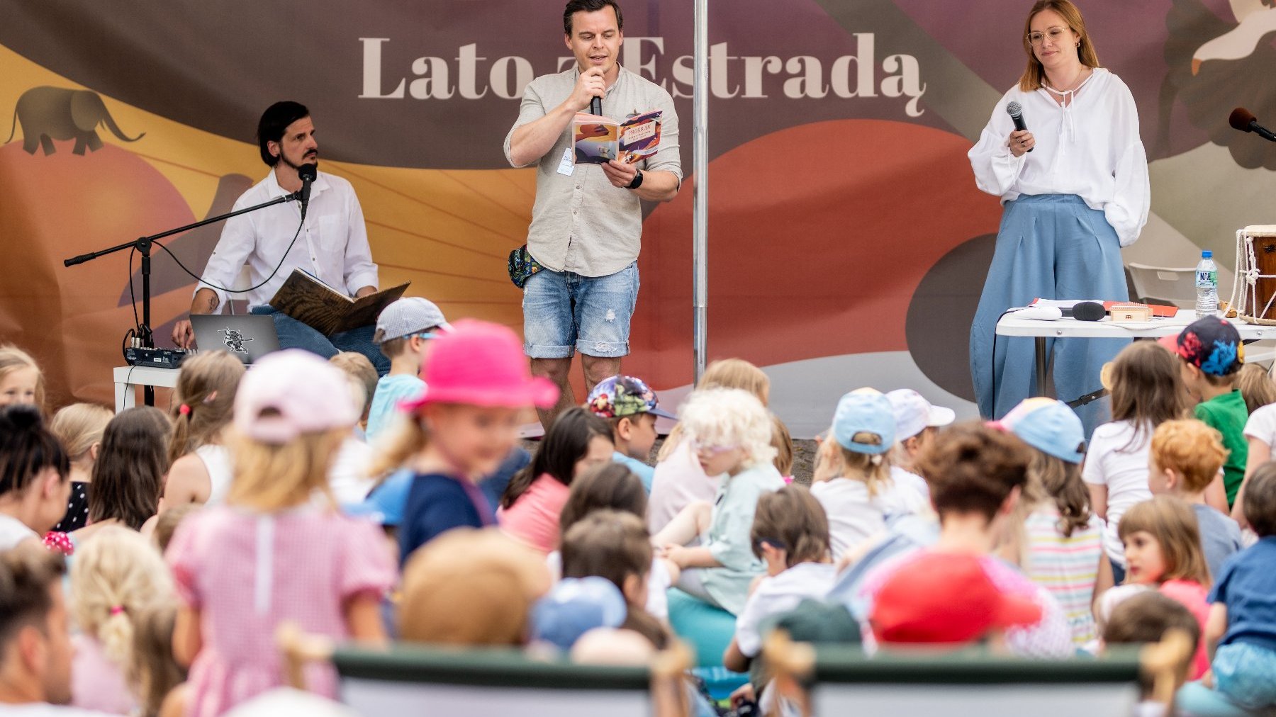 Na zdjęciu trójka na ludzi na scenie, przed nimi tłum dzieci oraz młodzieży