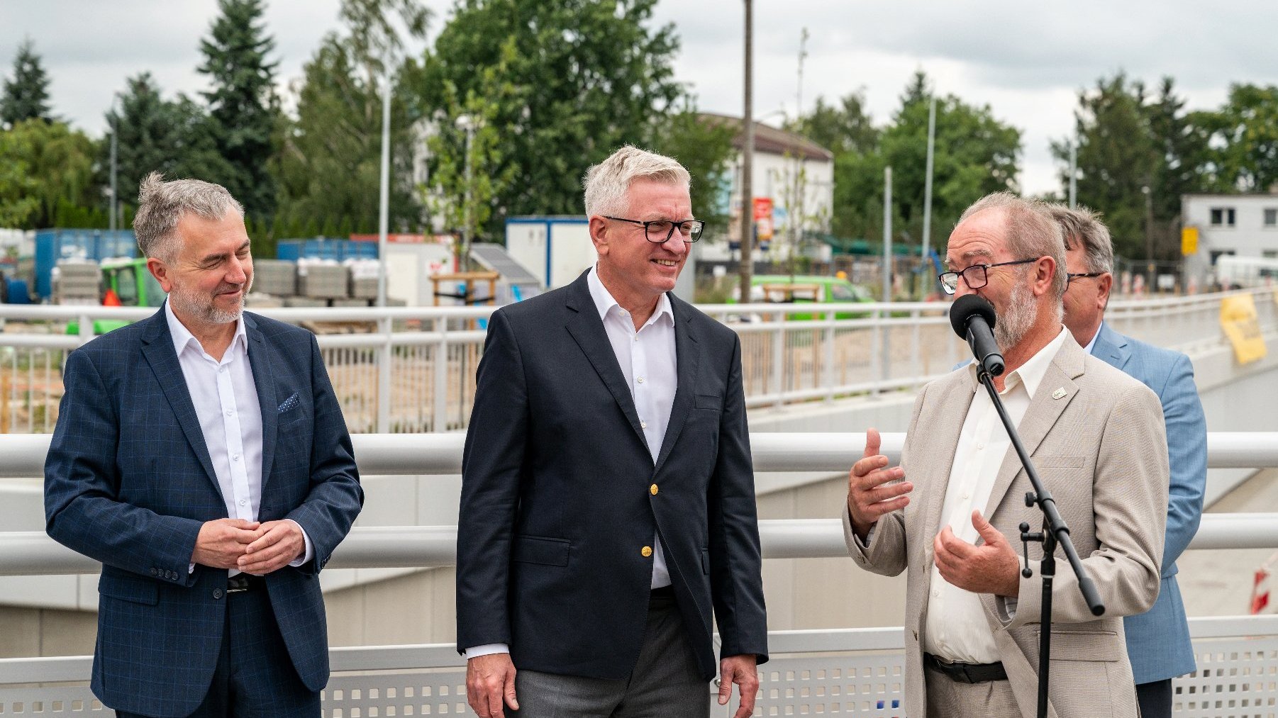 Na zdjeciu czterech mężczyzn w garniturach, jeden z nich przy mikrofonie