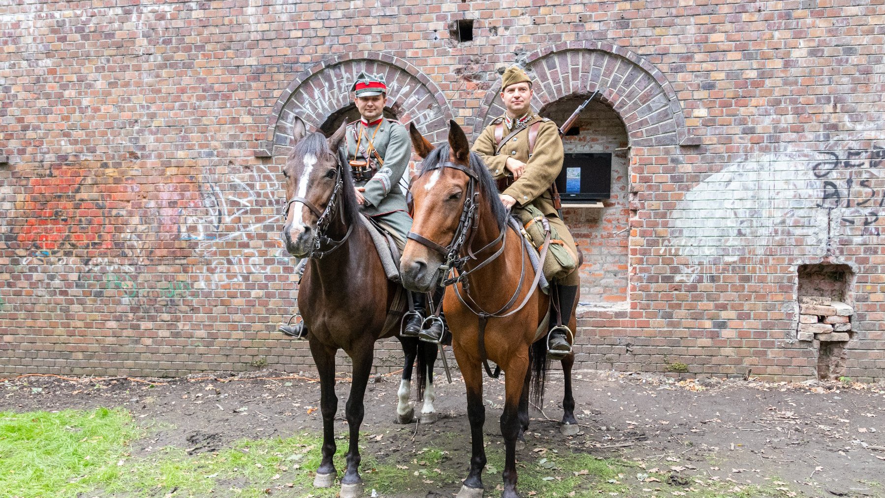 Na zdjęciu dwóch rekonstruktorów w mundurach, na koniach, przed fortem