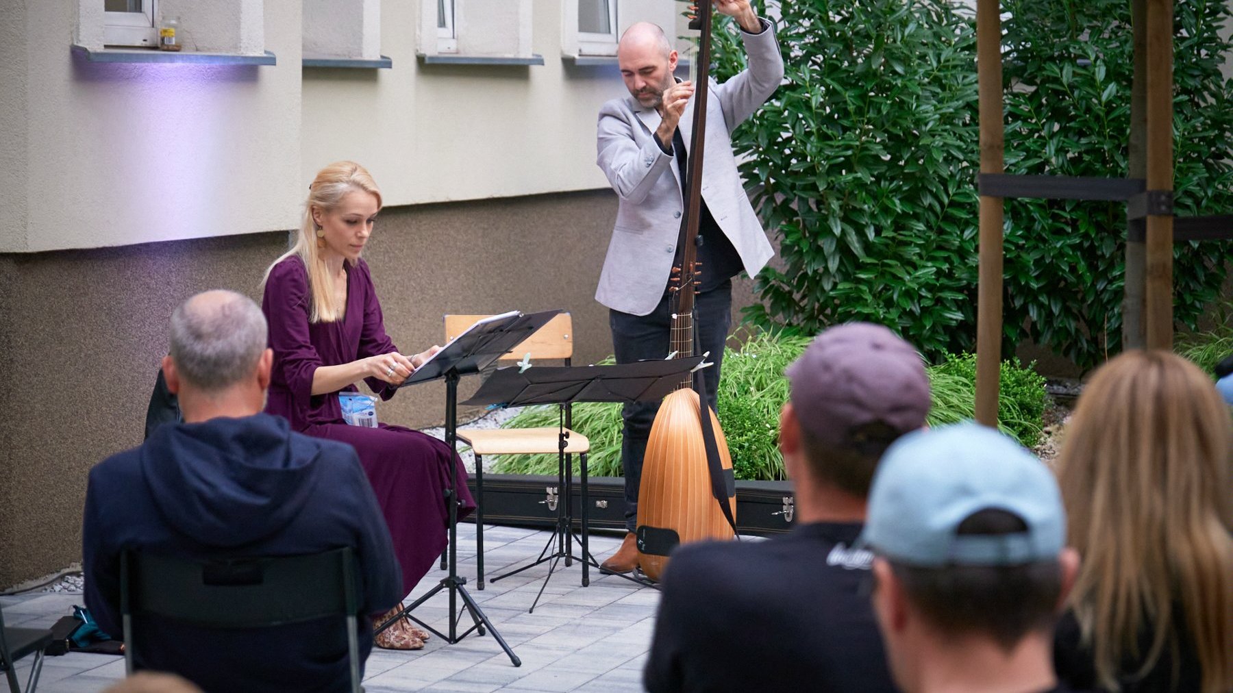 Na zdjęciu kobieta siedzi przed stojakiem z nutami, obok niej mężczyzna gra na instrumencie, przed nimi siedzą widzowie