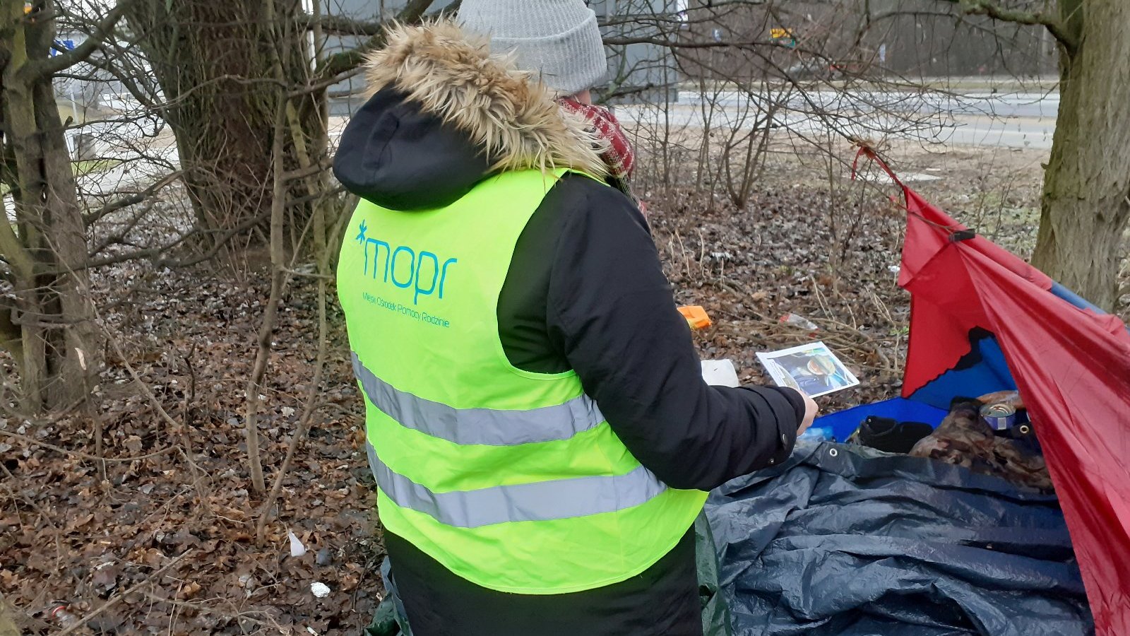 Na zdjęciu pracownica socjalna w kamizelce MOPR, stoi przy namiocie osoby w kryzysie bezdomności